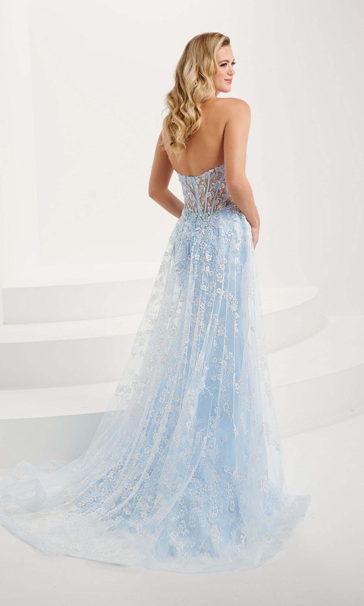 A woman models the Tiffany Strapless Sheer Corset Prom Dress 16107, featuring a pastel hue, sheer overlay, and floral sequin embroidery. She stands on a white background, facing away and glancing over her shoulder.